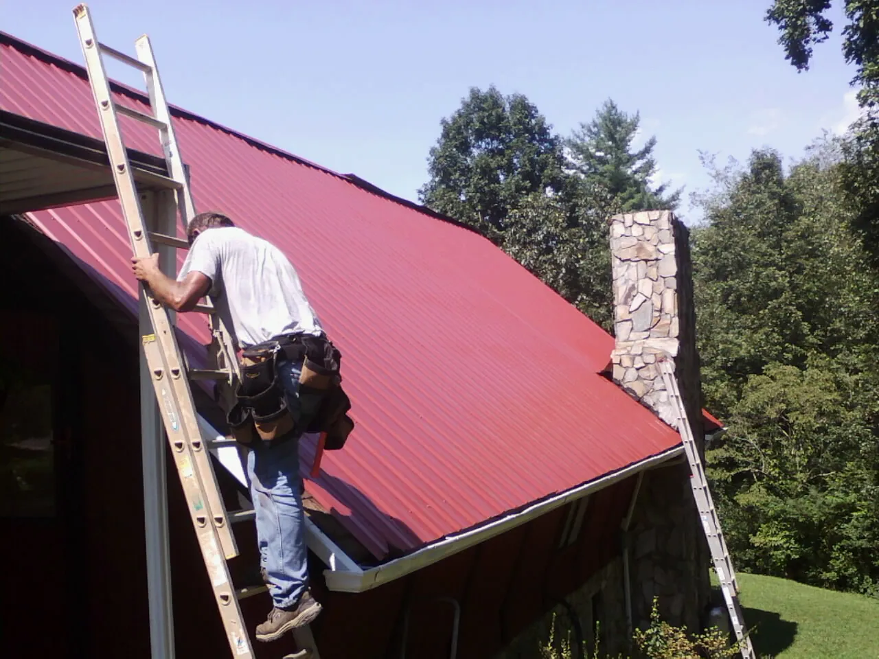 Roofer on a ladder beside a red metal roof and stone chimney — Soffit & Fascia Repair by Roofing Brothers in East Texas